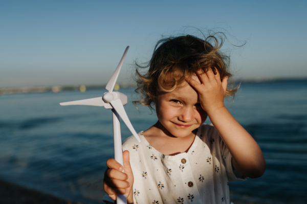 Small girl with blonde hair standing at beach, holding wind turbine model in the hand, sea wind spinning blades.