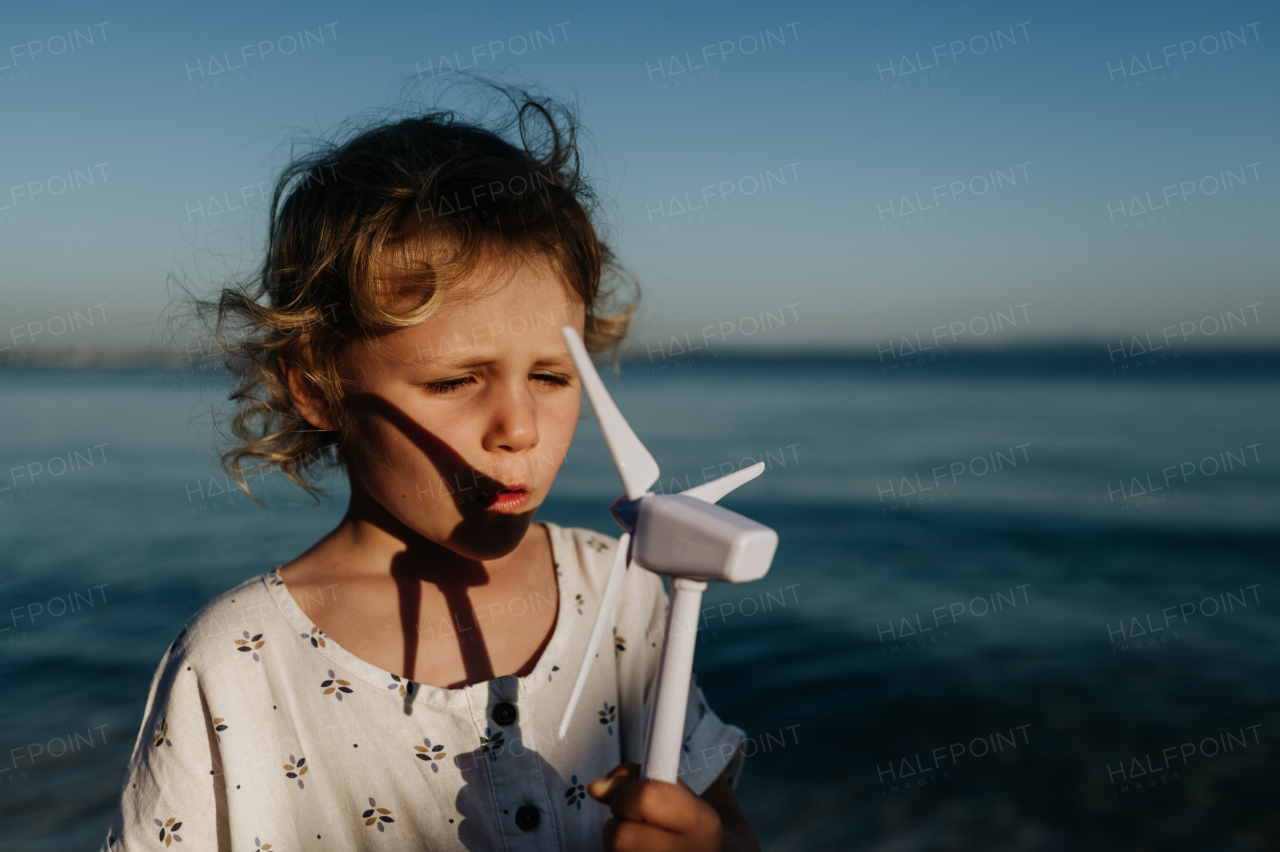 Small girl with blonde hair standing at beach, holding wind turbine model in the hand, sea wind spinning blades.