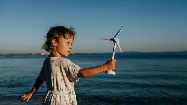 Small girl with blonde hair standing at beach, holding wind turbine model in the hand, sea wind spinning blades.