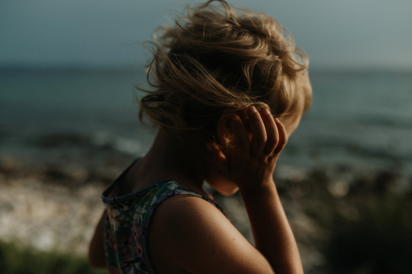 Side view of small girl with blonde hair standing at the beach looking at sea.