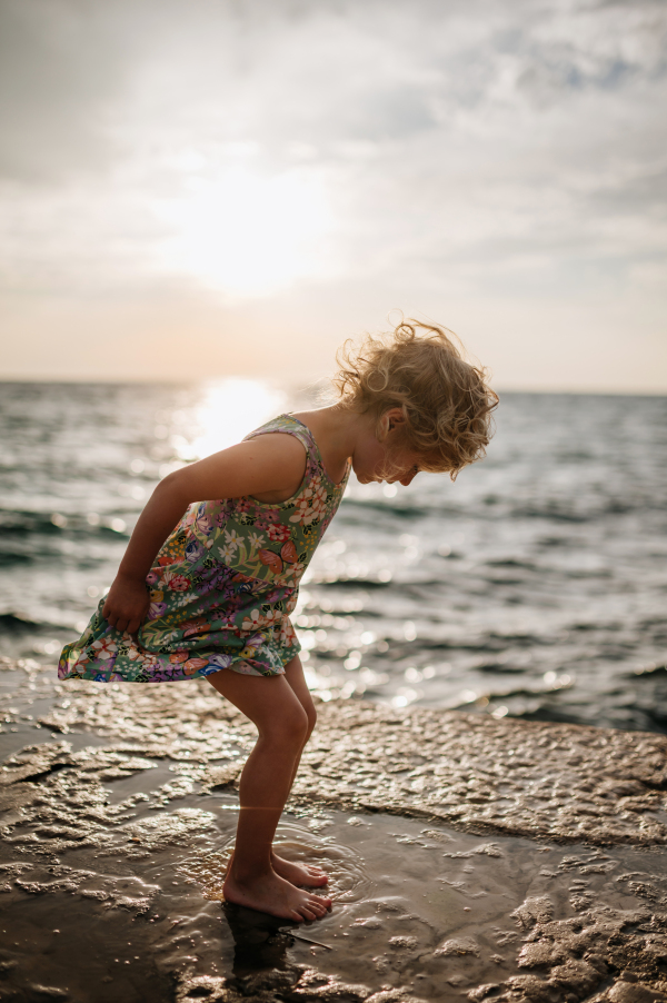 Young girl jumping in puddle on promenade, seawall by sea on sand beach. Barefoot happy girl in summer dress having fun.