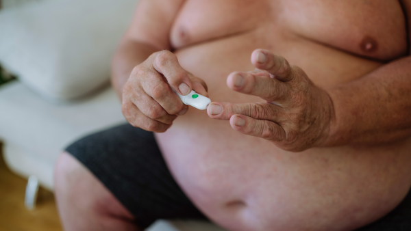 Overweight man checking blood glucose level at home using a fingerstick glucose meter. The diabetic man taking a finger-prick blood sample.