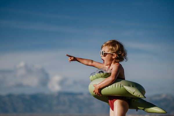 Side view of small surprised girl at beach with inflatable toy ring, pointing with finger into distance.