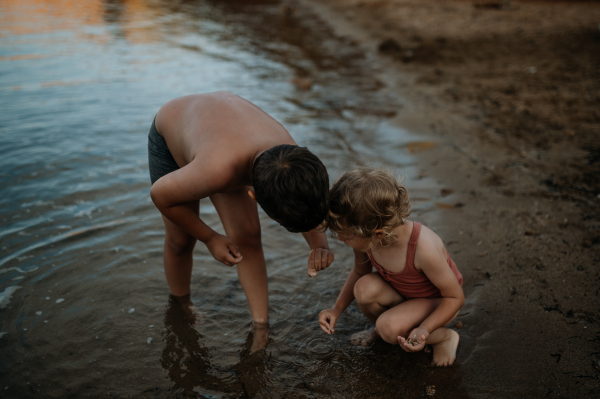 Brother helping sister seraching for shells in wet sand. Small girl in swimsuit playing at beach, crouching in water.