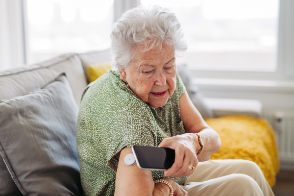 Diabetic senior patient checking blood glucose level at home using continuous glucose monitor. Elderly woman connecting her CGM with smarphone to see her blood sugar levels in real time.