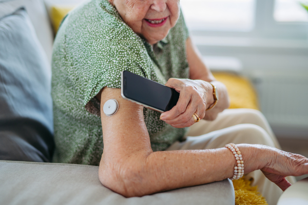 Close up of diabetic senior patient checking blood glucose level at home using continuous glucose monitor. Elderly woman connecting her CGM with smarphone to see her blood sugar levels in real time.