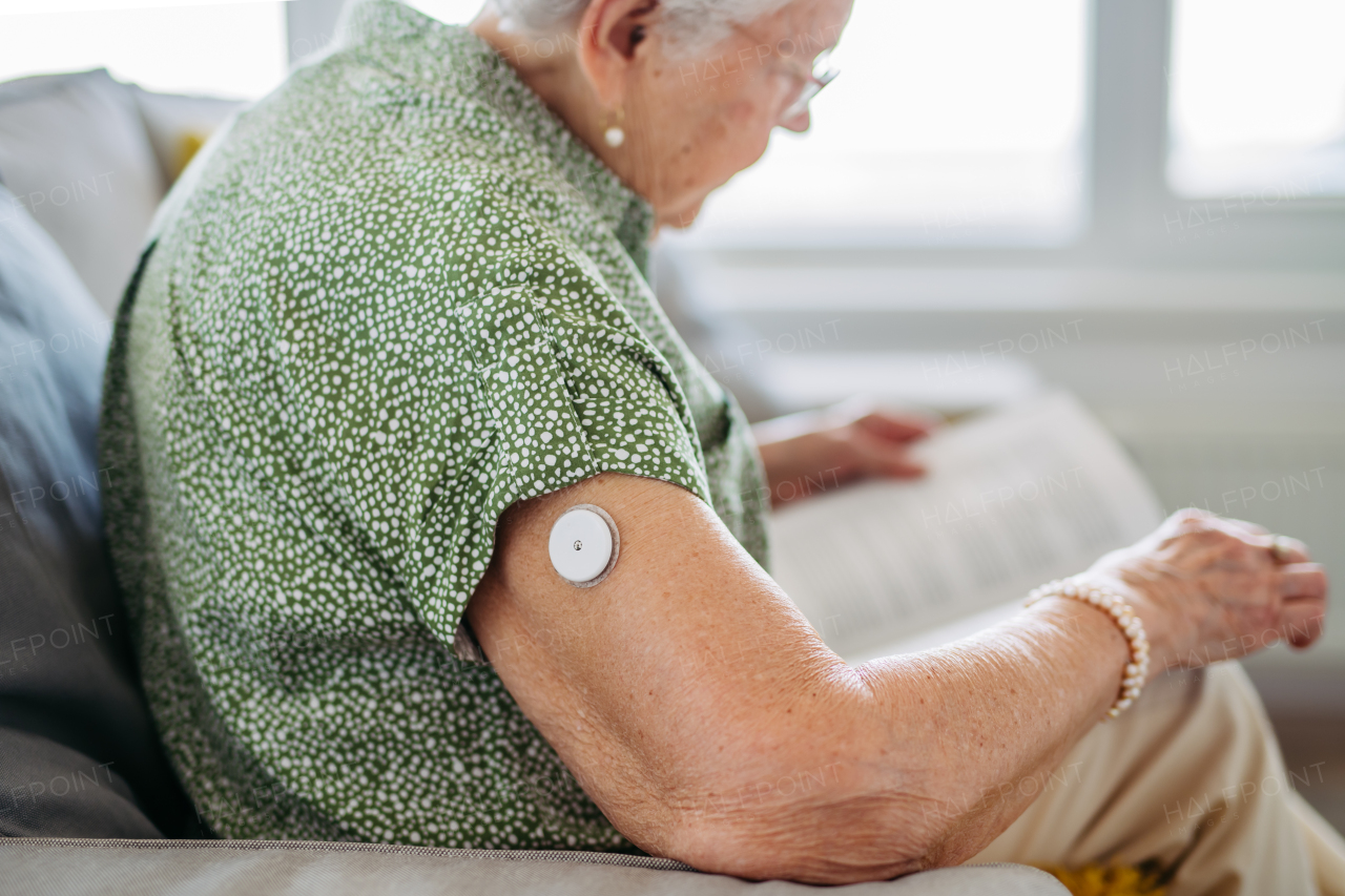 Diabetic senior patient using continuous glucose monitor to check blood sugar level at home. CGM device making life of elderly woman easier, helping manage her illness and focus on other activities. Senior diabetic woman reading book at home.