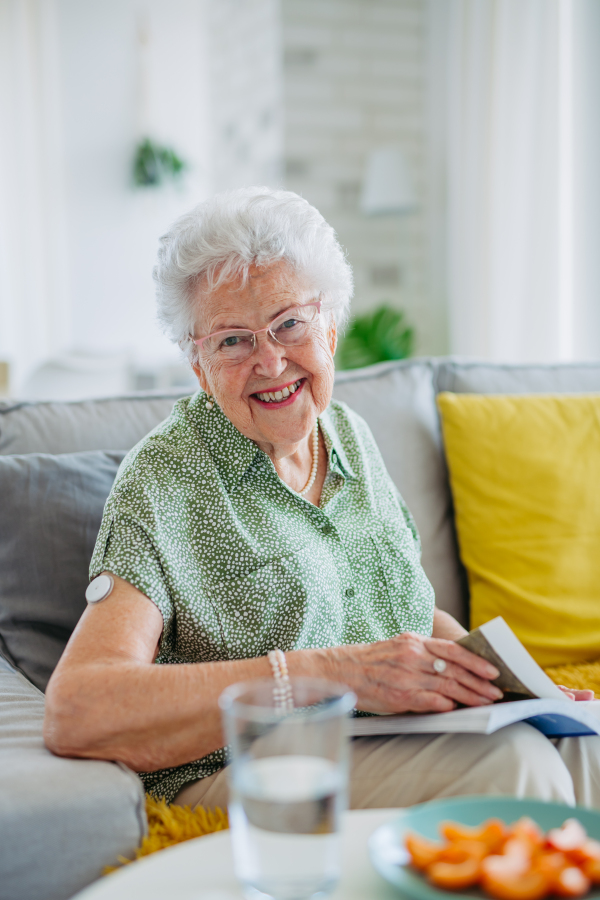 Diabetic senior patient using continuous glucose monitor to check blood sugar level at home. Senior woman drinking water to better manage her diabetes. CGM device making life of elderly woman easier. Old lady reading at home.