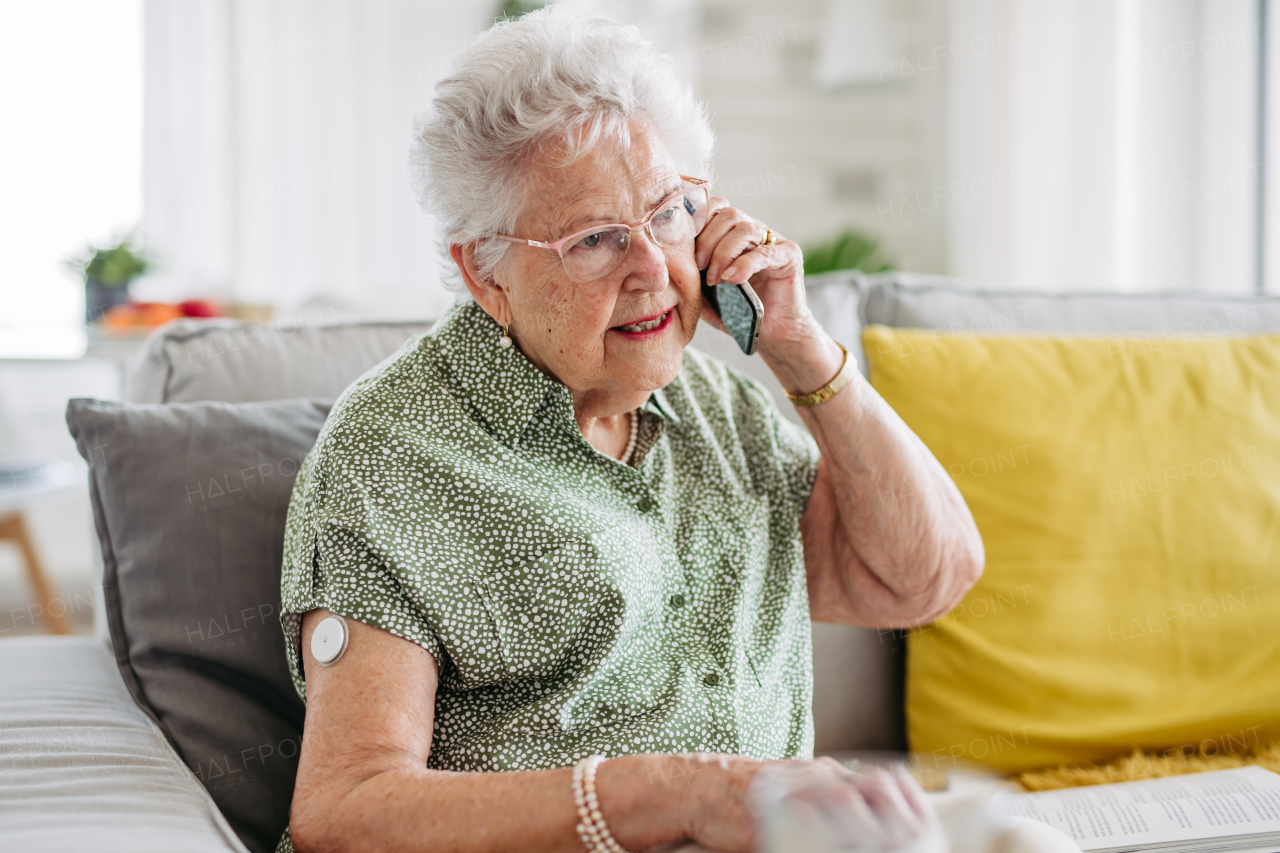 Diabetic senior patient using continuous glucose monitor to check blood sugar level at home. CGM device making life of elderly woman easier, helping manage her illness and focus on other activities. Modern senior diabetic woman making call on her smartphone.