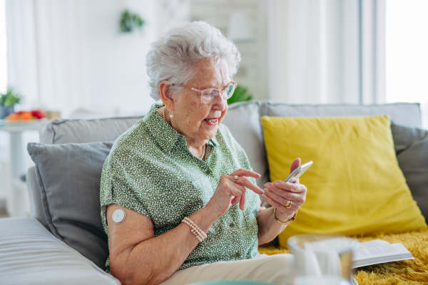 Diabetic senior patient using continuous glucose monitor to check blood sugar level at home. CGM device making life of elderly woman easier, helping manage her illness and focus on other activities. Senior diabetic woman checking her glucose data on smartphone.