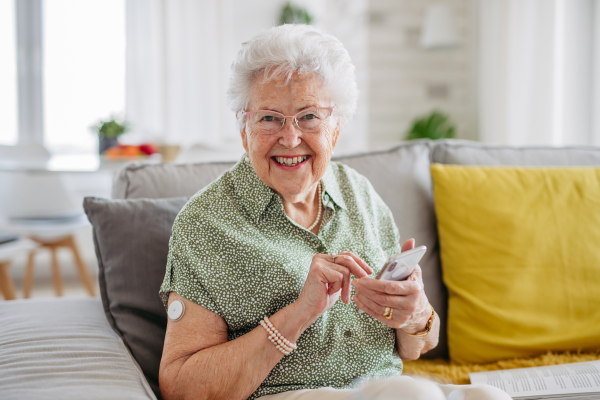 Diabetic senior patient using continuous glucose monitor to check blood sugar level at home. CGM device making life of elderly woman easier, helping manage her illness and focus on other activities. Senior diabetic woman checking her glucose data on smartphone.