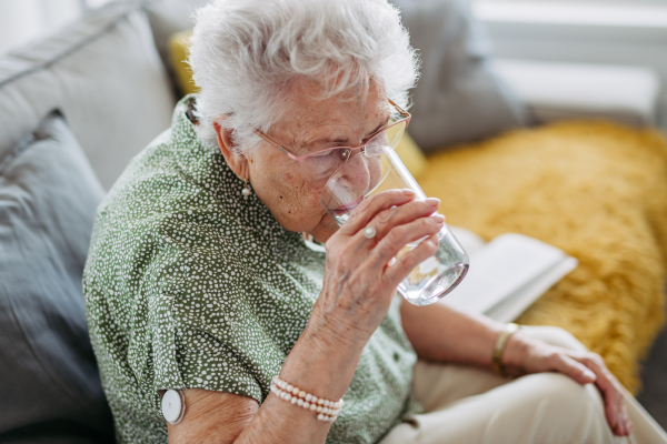 Diabetic senior patient using continuous glucose monitor to check blood sugar level at home. Senior woman drinking water to better manage her diabetes. CGM device making life of elderly woman easier.