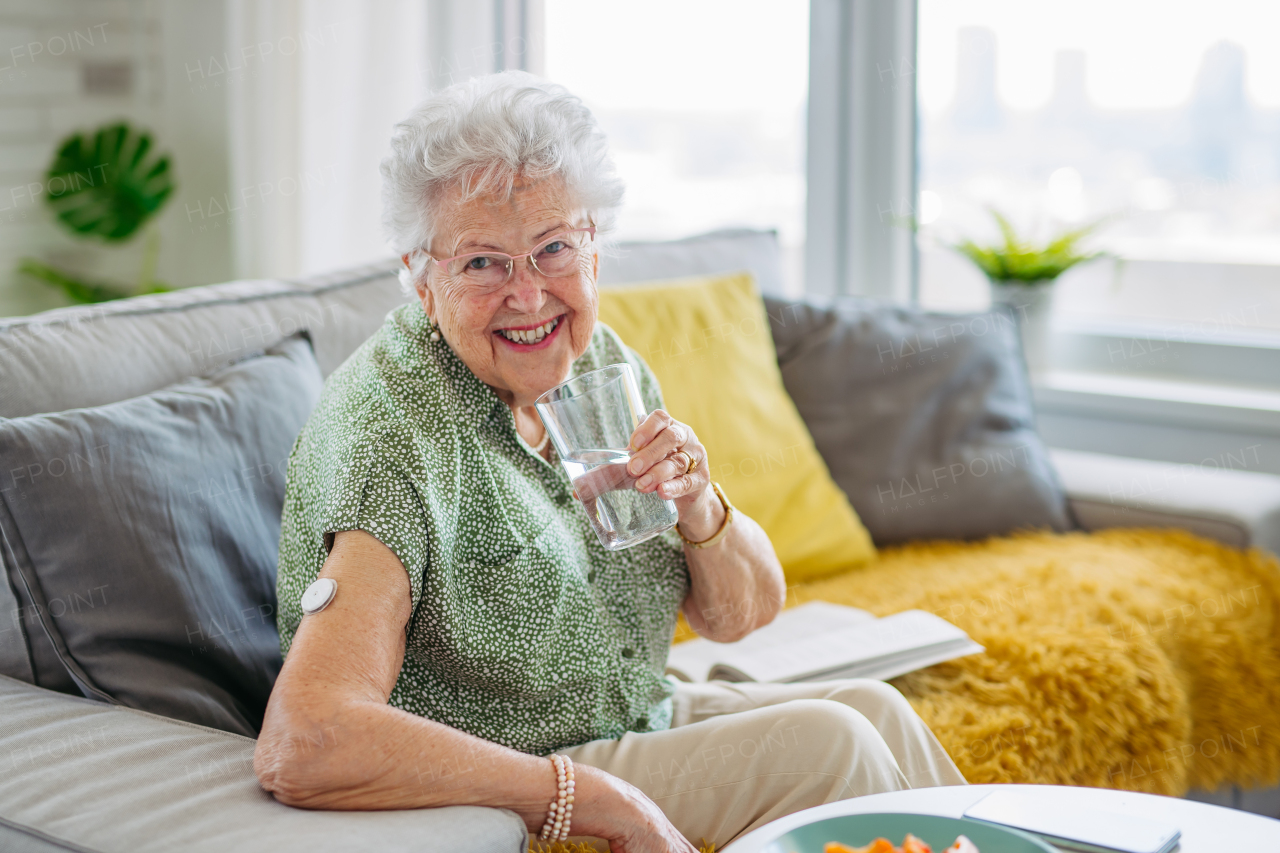 Diabetic senior patient using continuous glucose monitor to check blood sugar level at home. Senior woman drinking water to better manage her diabetes. CGM device making life of elderly woman easier.