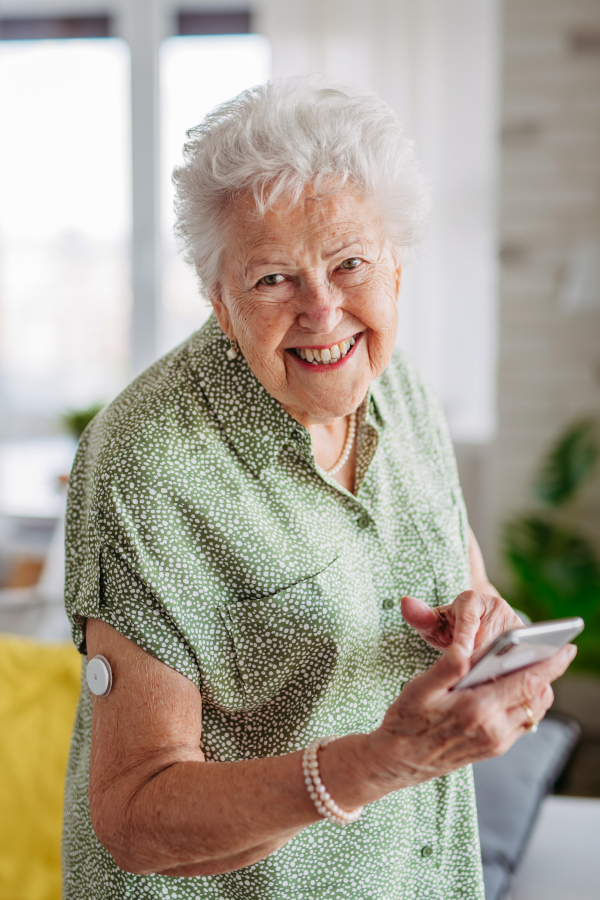 Senior diabetic woman checking her glucose data on smartphone. Smiling elderly lady holding mobile phone. Diabetic senior patient using continuous glucose monitor to check blood sugar level at home.