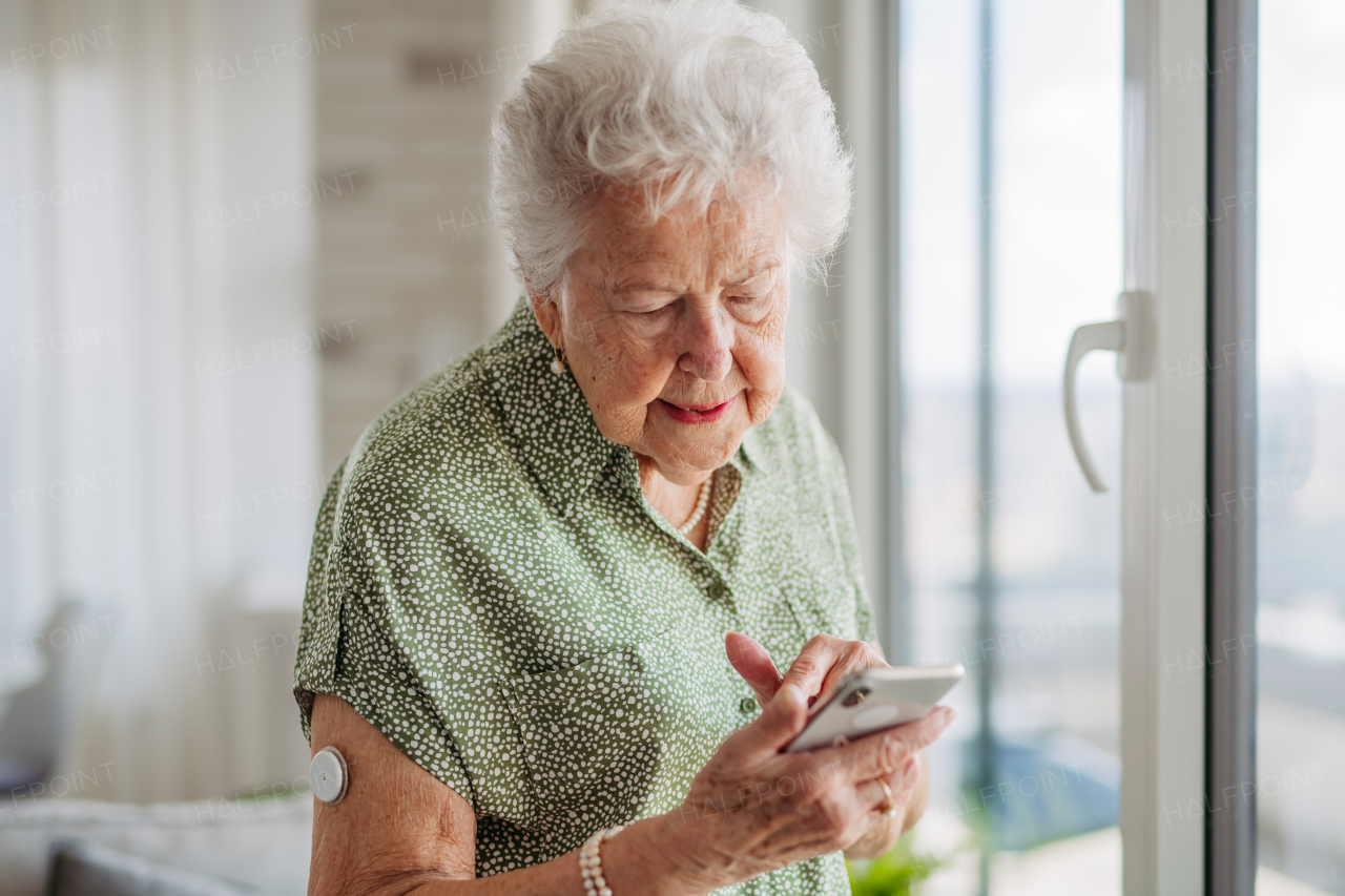 Senior diabetic woman checking her glucose data on mobile phone. Smiling elderly lady scrolling on smart phone. Diabetic senior patient using continuous glucose monitor to check blood sugar level at home.