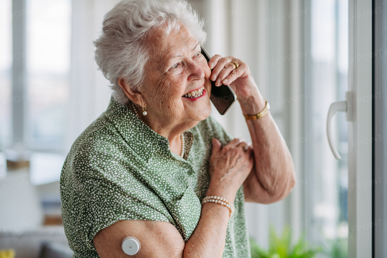 Diabetic senior patient using continuous glucose monitor to check blood sugar level at home. CGM device making life of elderly woman easier, helping manage her illness and focus on other activities. Senior diabetic woman making phone call at home.