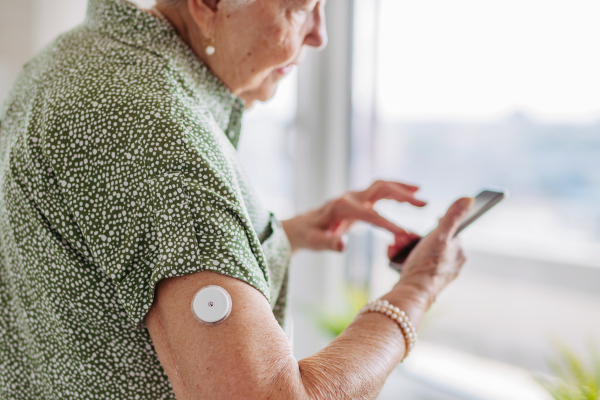 Diabetic senior patient using continuous glucose monitor to check blood sugar level at home. CGM device making life of elderly woman easier, helping manage her illness and focus on other activities. Senior diabetic woman checking her glucose data on smartphone.