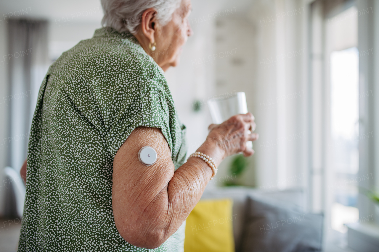 Diabetic senior patient using continuous glucose monitor to check blood sugar level at home. Senior woman drinking water to better manage her diabetes. CGM device making life of elderly woman easier.