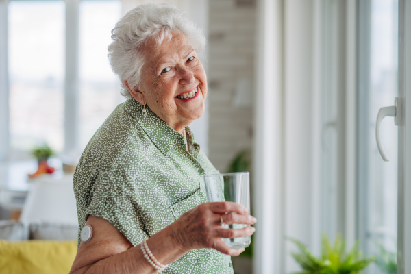 Diabetic senior patient using continuous glucose monitor to check blood sugar level at home. Senior woman drinking water to better manage her diabetes. CGM device making life of elderly woman easier.