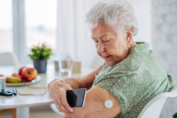 Diabetic senior patient checking blood glucose level at home using continuous glucose monitor. Elderly woman connecting her CGM with smarphone to see her blood sugar levels in real time.