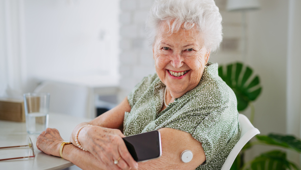 Diabetic senior patient checking blood glucose level at home using continuous glucose monitor. Elderly woman connecting her CGM with smarphone to see her blood sugar levels in real time. Banner with copy space.