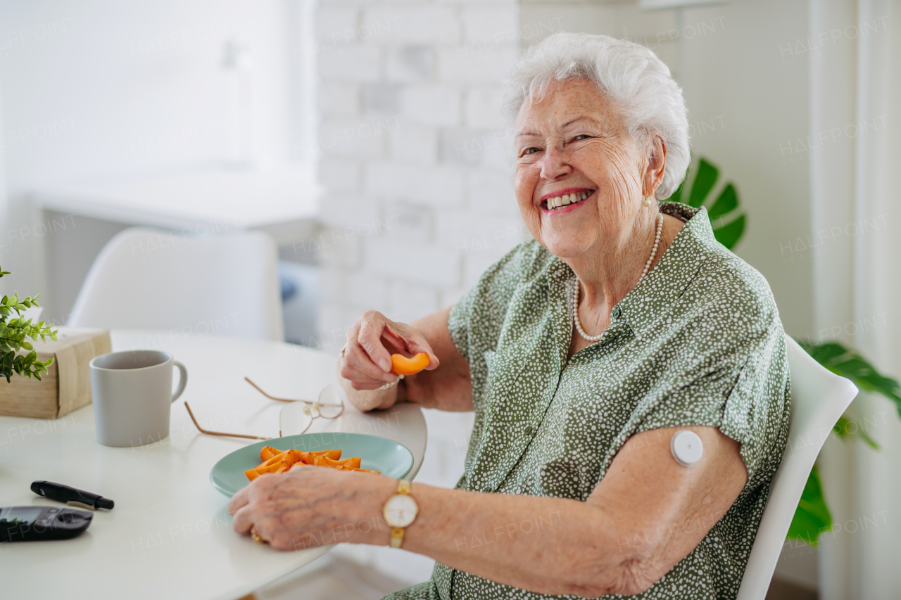 Diabetic senior patient using continuous glucose monitor to check blood sugar level at home. Senior woman eating fruit to help raise her blood sugar to normal. CGM device making life of elderly woman easier, helping manage her illness and focus on other activities.