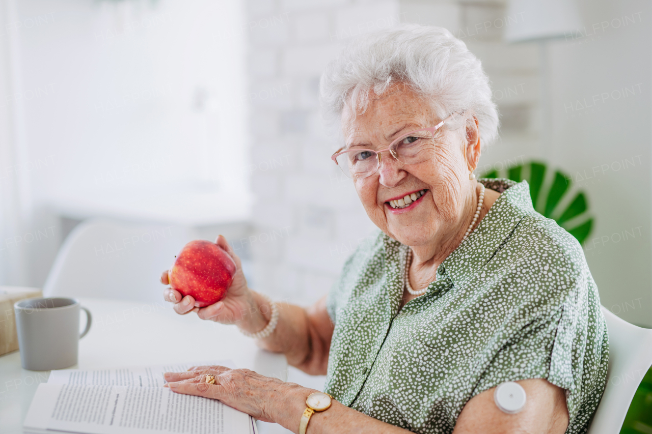 Diabetic senior patient using continuous glucose monitor to check blood sugar level at home. Senior woman eating apple to help raise her blood sugar to normal. CGM device making life of elderly woman easier, helping manage her illness and focus on other activities.