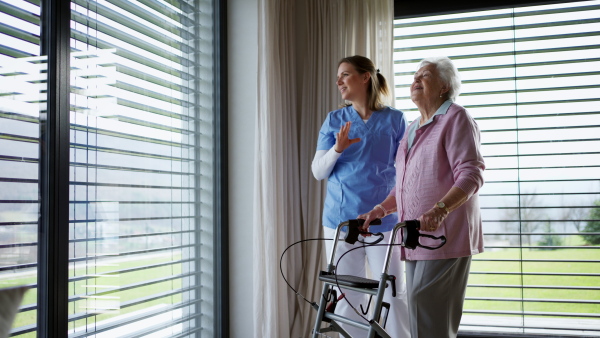 Caregiver, nurse helping senior woman move around the house, looking out the window, enjoying the view. Elderly woman walking indoors, using walker with wheels.