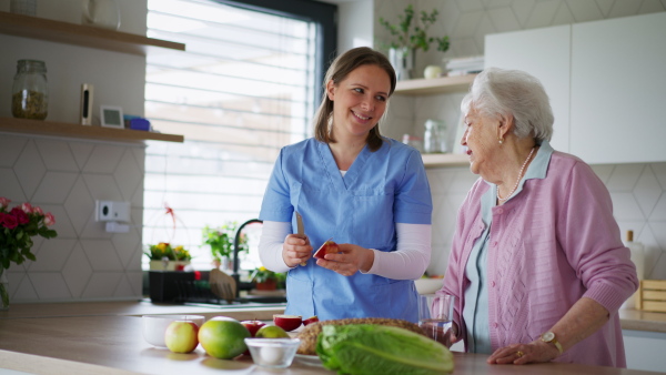 Home caregiver and elderly woman cutting apples, making healthy quick snack. Household chores for senior woman, help from nurse.