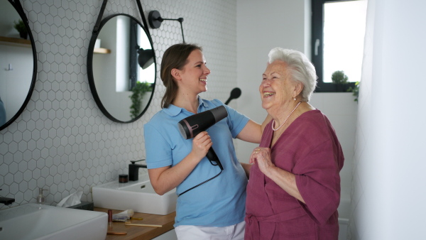 Home caregiver helping elderly woman in bathroom, combing her hair. Senior woman at home with young beautiful nurse.