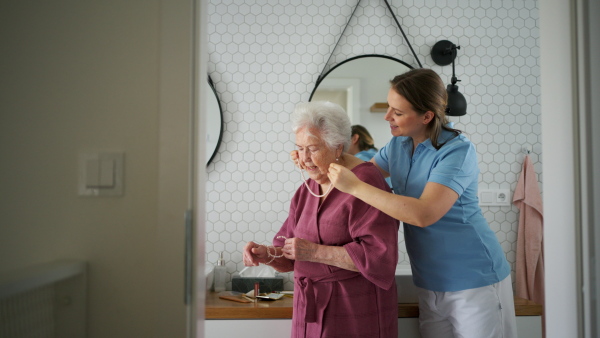 Home caregiver helping elderly woman in bathroom, putting on jewellery. Senior woman at home with young beautiful nurse