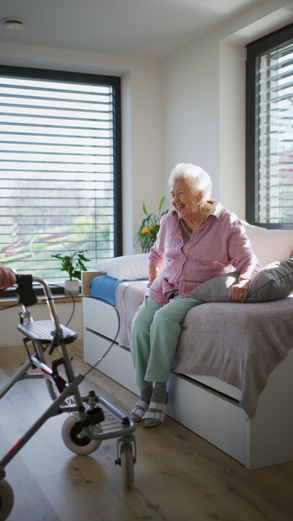 Caregiver, nurse helping senior woman to stand from bed in home, waiting patiently. Elderly woman sitting on bed, trying to get up, smiling.