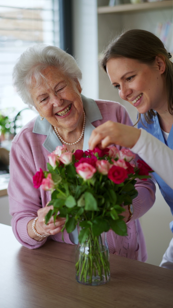 Elderly woman receiving bouquet of roses to her female home caregiver. Senior woman at home is thankful for care and assistance from nurse.