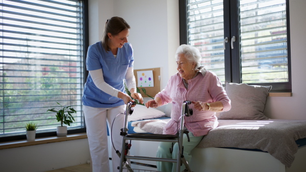 Caregiver, nurse helping senior woman to stand from bed in home, waiting patiently. Elderly woman sitting on bed, trying to get up, smiling.