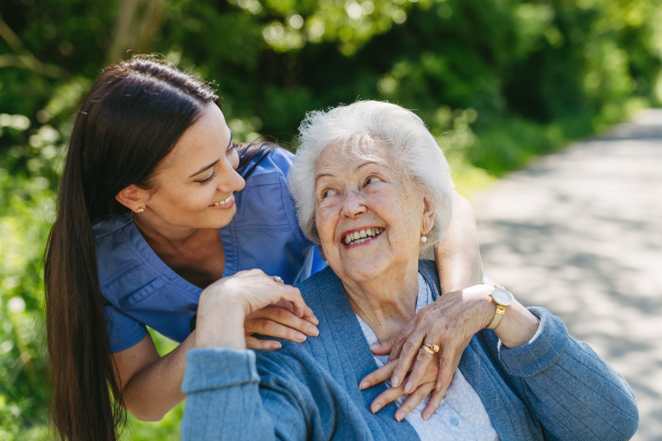 Caregiver pushing senior woman in wheelchair. Nurse and elderly woman enjoying a warm day in nursing home, public park.