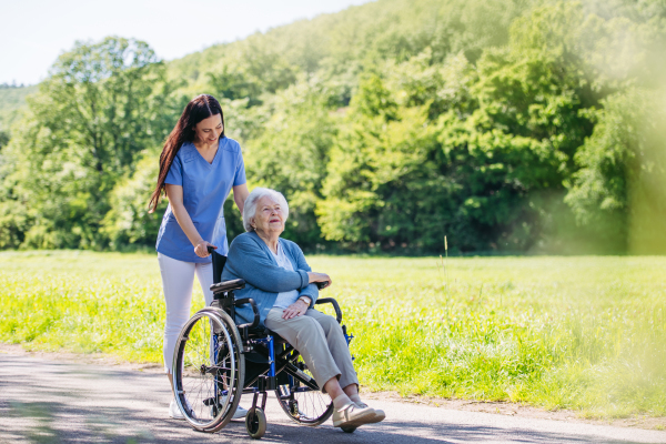 Caregiver pushing senior woman in wheelchair. Nurse and elderly woman enjoying a warm day in nursing home, public park.