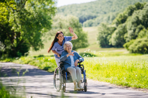 Caregiver pushing senior woman in wheelchair. Nurse and elderly woman enjoying a warm day in nursing home, public park.