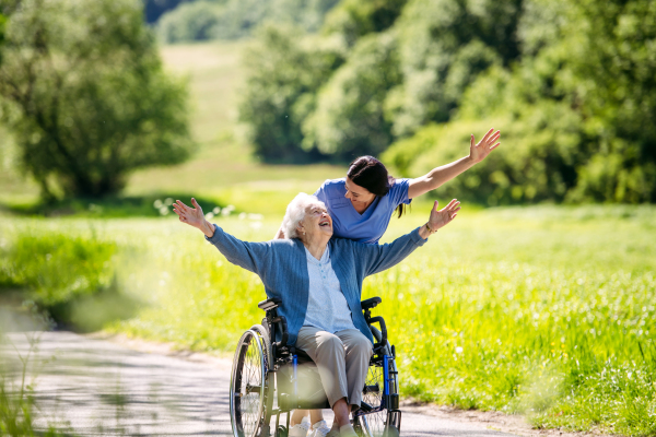 Caregiver pushing senior woman in wheelchair. Nurse and elderly woman enjoying a warm day in nursing home, public park.