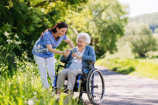 Caregiver and senior woman in wheelchair picking wild flowers. Nurse and elderly woman enjoying a warm day in nursing home, public park.