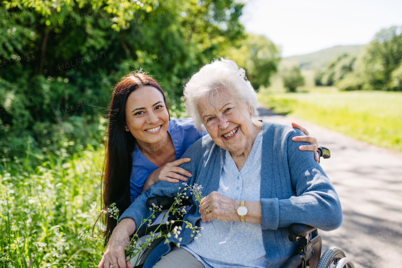 Caregiver and senior woman in wheelchair picking wild flowers. Nurse and elderly woman enjoying a warm day in nursing home, public park.