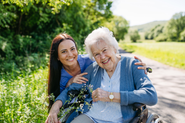 Caregiver and senior woman in wheelchair picking wild flowers. Nurse and elderly woman enjoying a warm day in nursing home, public park.