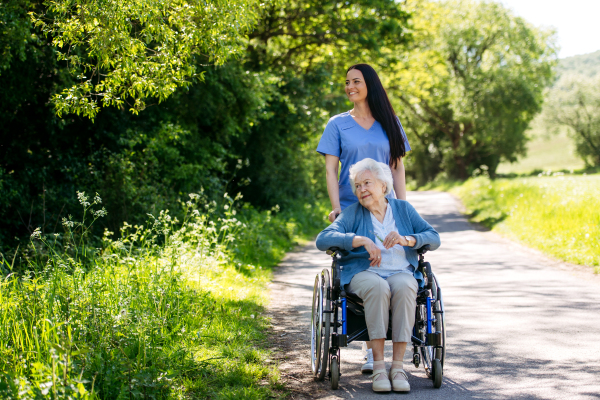 Caregiver pushing senior woman in wheelchair. Nurse and elderly woman enjoying a warm day in nursing home, public park.