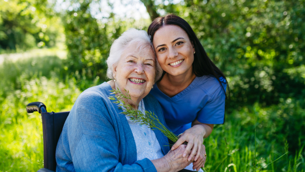 Female caregiver and senior woman in wheelchair. Nurse and elderly woman enjoying outdoor stroll during warm day in nature.