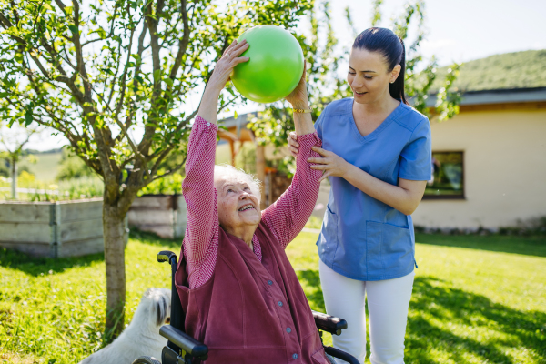 Female caregiver doing motorized exercises with senior woman in wheelchair. Nurse and elderly woman working out with green exercise ball.