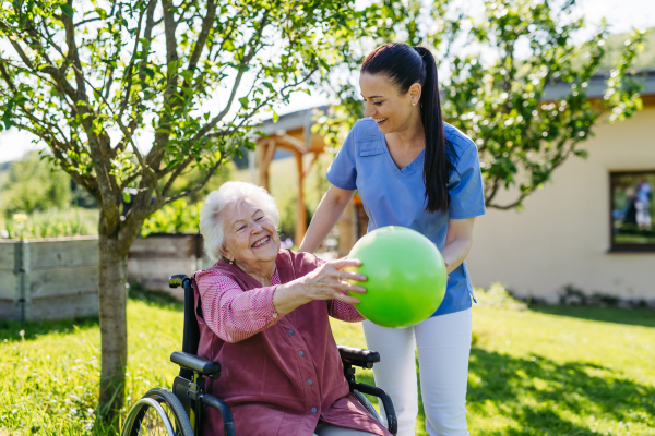 Female caregiver doing motorized exercises with senior woman in wheelchair. Nurse and elderly woman working out with green exercise ball.