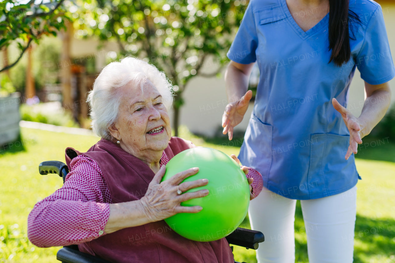 Female caregiver doing motorized exercises with senior woman in wheelchair. Nurse and elderly woman working out with green exercise ball.