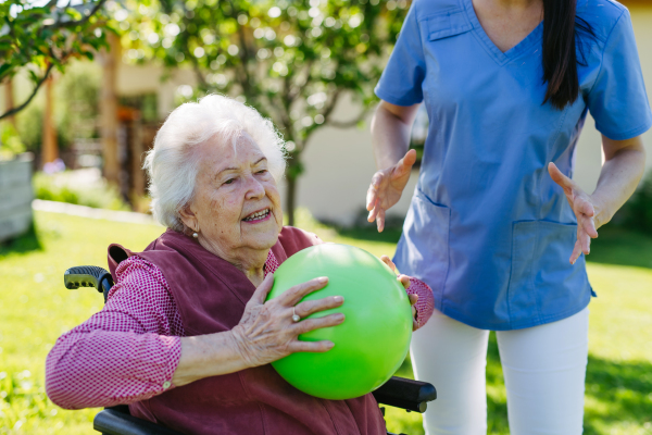 Female caregiver doing motorized exercises with senior woman in wheelchair. Nurse and elderly woman working out with green exercise ball.