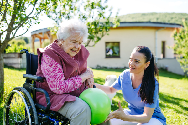 Female caregiver doing motorized exercises with senior woman in wheelchair. Nurse and elderly woman working out with green exercise ball.