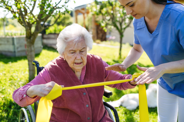 Female caregiver doing motorized exercises with senior woman in wheelchair. Nurse and elderly woman working out with yellow resistance band.