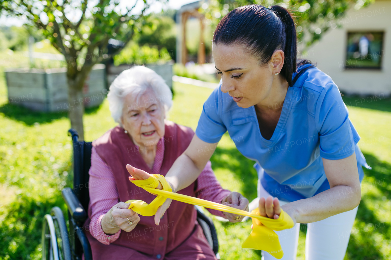 Female caregiver doing motorized exercises with senior woman in wheelchair. Nurse and elderly woman working out with yellow resistance band.